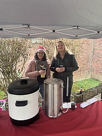Zwei Frauen stehen hinter einem Tisch mit zwei Wärmebehältern und haben jeweils einen Becher in der Hand.