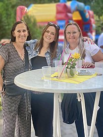 Drei Frauen gemischten Alters stehen an einem weißen Stehtisch und tragen sommerliche Kleidung.