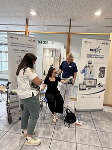 Eine Frau sitzt in einem medizinischen Stuhl und wird auf die Funktionsweise des Gerätes aufmerksam gemacht durch eine weitere Frau in Funktionskleidung. Eine dritte Frau schaut zu.