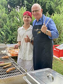Ein junger Teenager mit roter Cap und ein älterer Mann mit Schürze und Grillzange in der Hand stehen vor einem großen Grill.