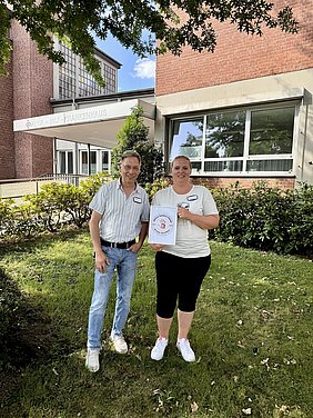 Ein Mann und eine Frau mit einem Zertifikat in der Hand auf einem Grünstreifen vor einem Krankenhauseingang