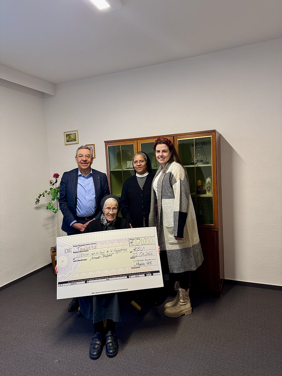 Gruppenbild von einem älteren mann im Anzug hinten, einer indischen Ordensschwester hinten mitte und einer jungen Frau hinten rechts sowie einer Ordensschwester sitzend vorne mit einem großen Spendenscheck in den Händen