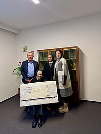 Gruppenbild von einem älteren mann im Anzug hinten, einer indischen Ordensschwester hinten mitte und einer jungen Frau hinten rechts sowie einer Ordensschwester sitzend vorne mit einem großen Spendenscheck in den Händen