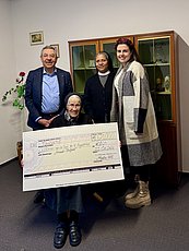 Gruppenbild von einem älteren mann im Anzug hinten, einer indischen Ordensschwester hinten mitte und einer jungen Frau hinten rechts sowie einer Ordensschwester sitzend vorne mit einem großen Spendenscheck in den Händen