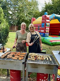 Zwei blonde Frauen (Tochter und Mutter) stehen an einem Grill im Garten, im Hintergrund eine Hüpfburg.