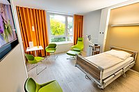 Renoviertes Einzelzimmer im Krankenhaus Maria-Hilf im Bergheim mit grünen Stühlen und Sessel, holzfarbenem hellem Bett, Schreibtisch und Nachttisch