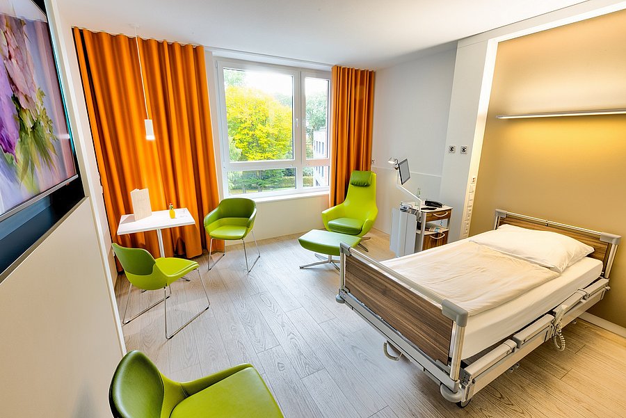 Renoviertes Einzelzimmer im Krankenhaus Maria-Hilf im Bergheim mit grünen Stühlen und Sessel, holzfarbenem hellem Bett, Schreibtisch und Nachttisch